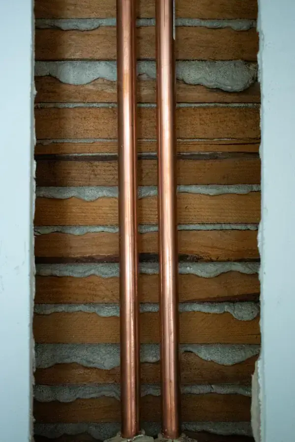 Two copper pipes run vertically through an exposed wall section, revealing wooden slats and gray plaster behind the pipes.
